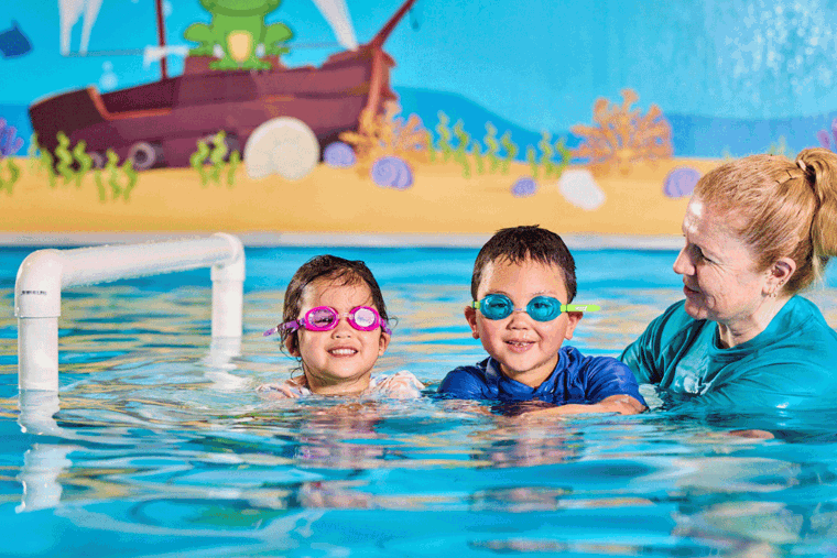 A woman with blonde hair is standing in a swimming pool in water is up to her shoulders. She's a swim instructor next to two children in the water with her. The children are preschool age, one is wearing pink goggles and one is wearing blue goggles.  Everyone is smiling and having fun.